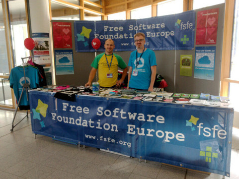 Two volunteers behind a booth of the FSFE presenting information and merchandise material.